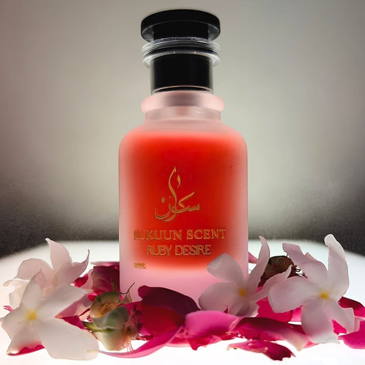 Sukuun Scent Ruby Desire Eau de Parfum 50ML, a vibrant red frosted glass bottle with gold lettering, surrounded by pink rose petals and white flowers.